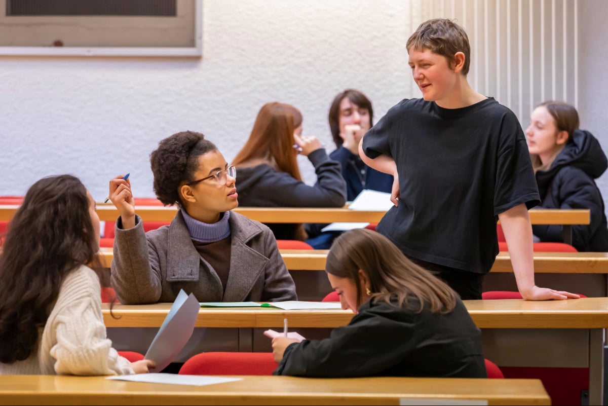 Students collaborating in a lecture hall, with a trainee solicitor standing and discussing with them while others listen, write notes, and hold worksheets.
