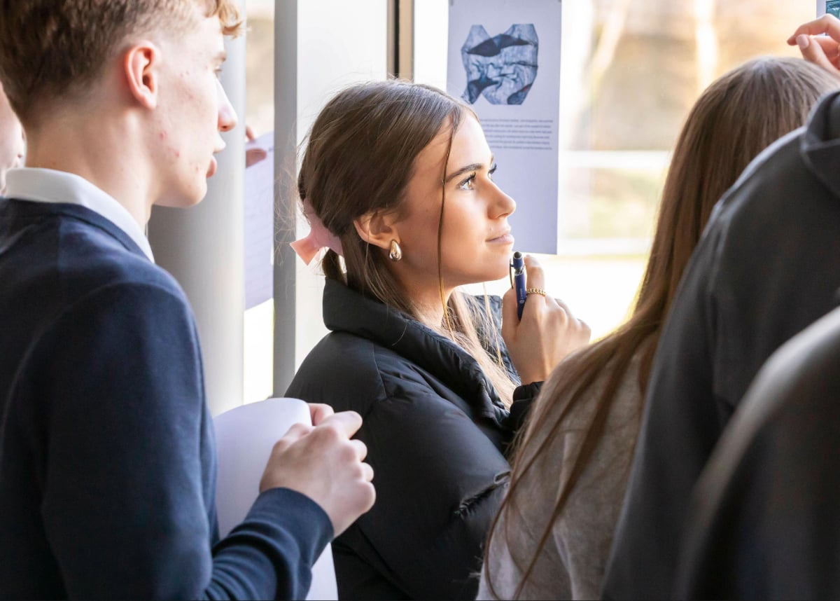 Students review and discuss a poster display near a window, with one student holding a pen and looking at the presentation material.