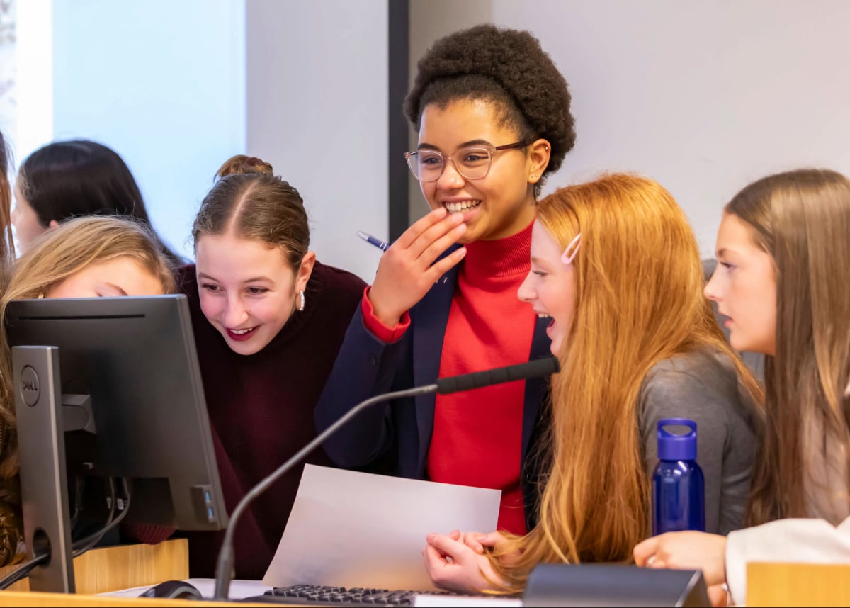 Students laughing and smiling together at a computer workstation, reviewing information on a monitor and holding printed materials.