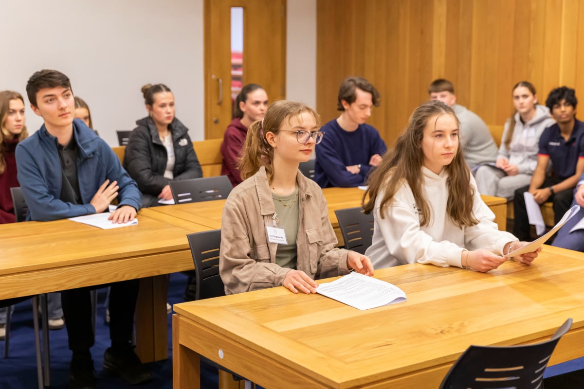 Students seated in a lecture room listening to a presentation, with papers on the desks and participants focused on the front of the room.