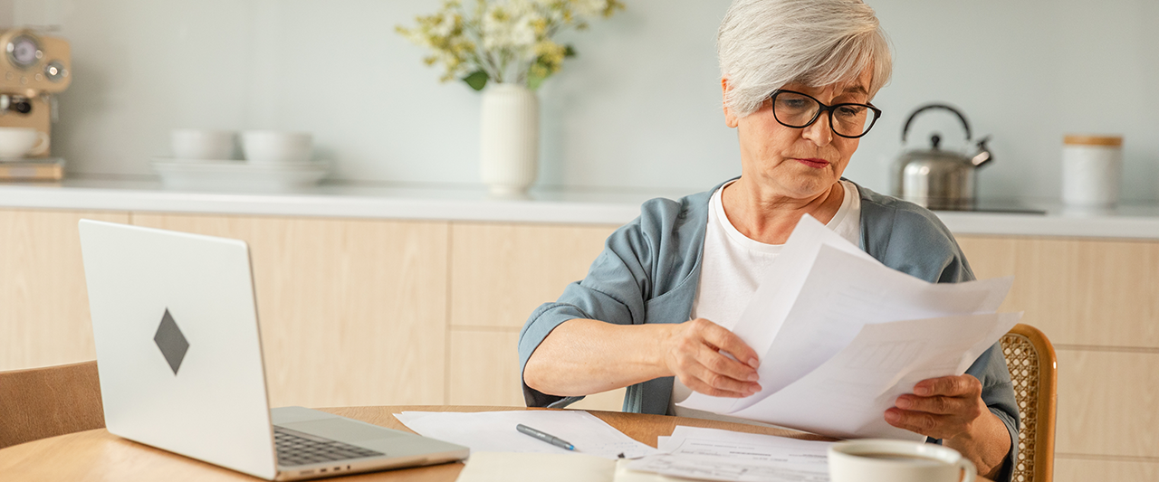 An older person looking at a document with a laptop computer in the background